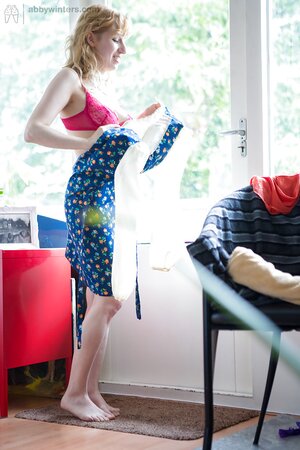 Teen in red bra and green panties wears a blue dress in front of a big window