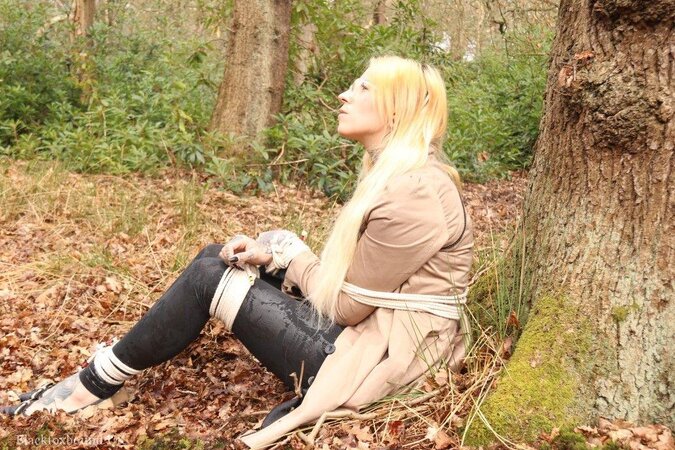 Girl with glasses can't stand up because she is hogtied by the tree